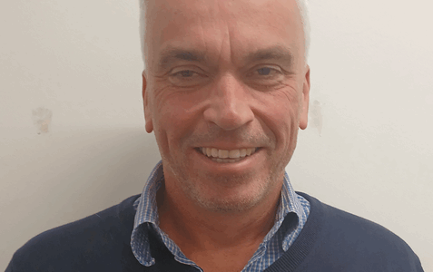 John Richardson ACS Finance Manager smiles to camera in blue jumper against white wall