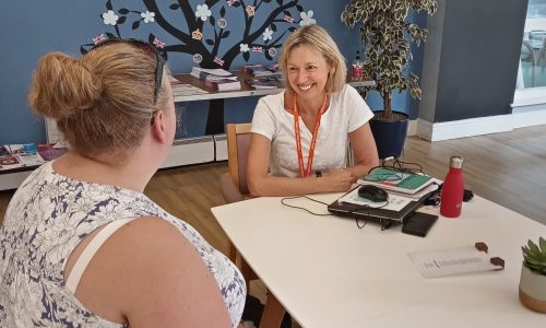 Two women are sat at a white table in a hall with blue wallpaper. The ACS Carer Supporter Adviser is smiling to the carer opposite as she offers help and information.