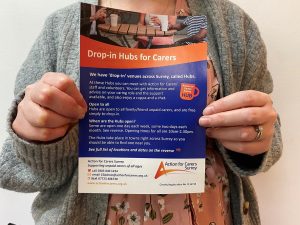 A woman's hands hold up a blue and orange leaflet which contains information about Action for Carers drop in Hubs. Woman wears orange dress and grey cardigan.