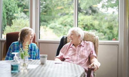 Older couple sit at table chatting with large window and garden view behind
