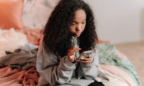 Teen girl sits on end of bed alone looking at phone thoughtful