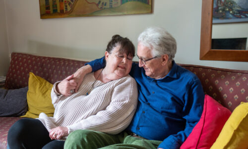 Older couple sat on sofa with man in blue jumper arm around shoulder of wife in white jumper, smiling