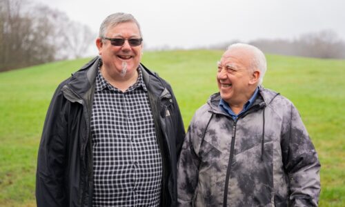 Two older men standing outside chatting and laughing with green grass background in winter